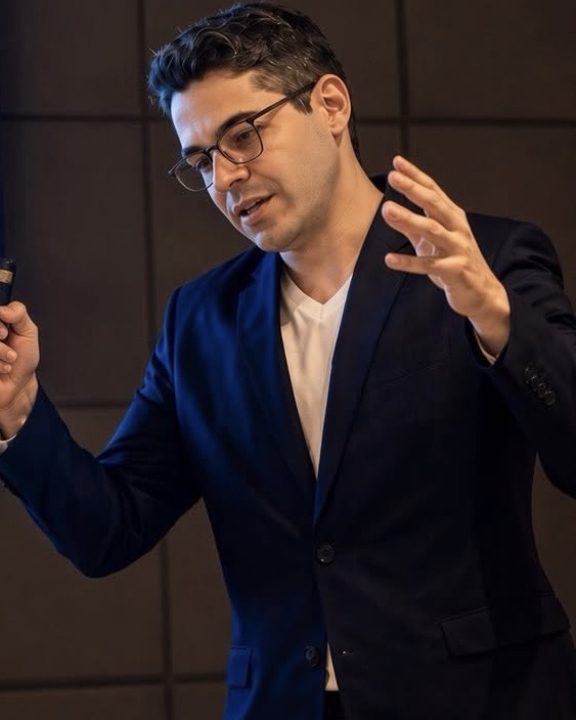 João Brígido em pé, de blazer escuro, explicando com as mãos durante uma aula