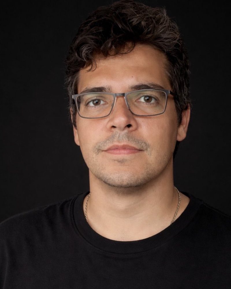 Retrato de João Brígido, de óculos e camiseta preta, sobre fundo escuro